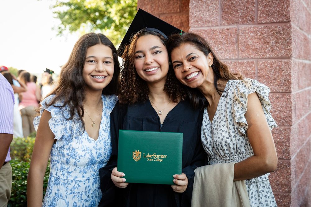 Three young women smile at graduation, one in cap and gown holding a Lake-Sumter State College diploma.