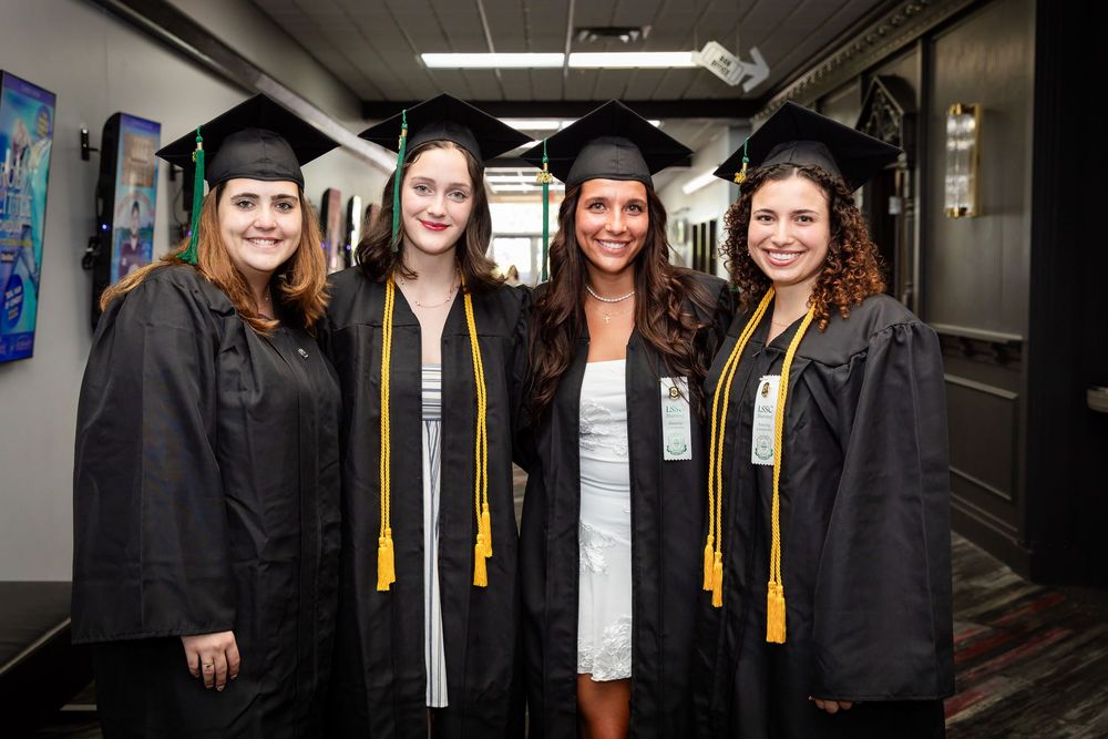 Four female graduates in black caps and gowns with gold tassels smile together at a graduation ceremony.