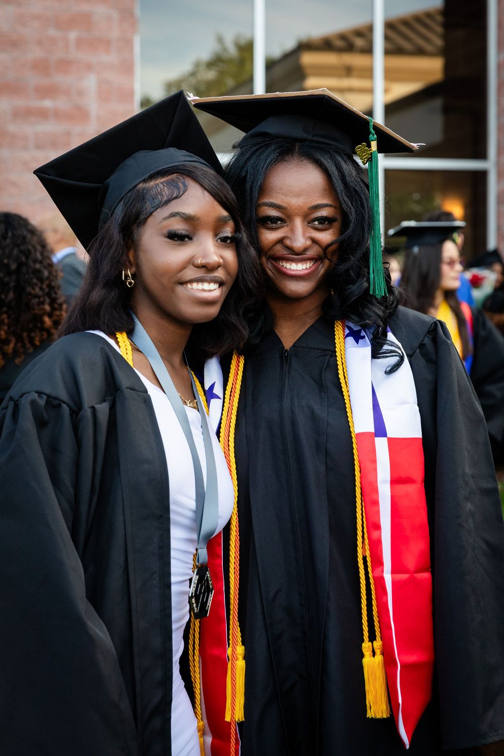 Two smiling women in black graduation caps and gowns with gold tassels and colorful stoles celebrate commencement.