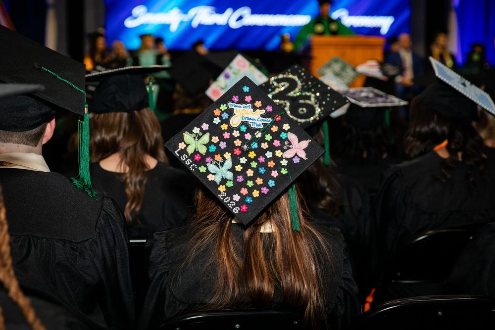 Graduates in black caps and gowns at a commencement ceremony, featuring a decorated cap with colorful dots and 2025.