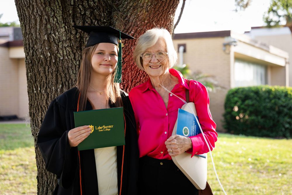 Young graduate in cap and gown holds diploma beside elderly woman near tree at outdoor graduation celebration.