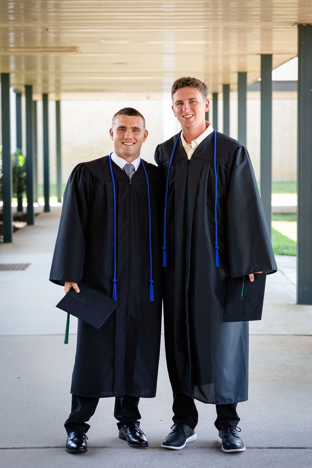 Two young men in black graduation gowns with blue cords smile proudly in a campus hallway.