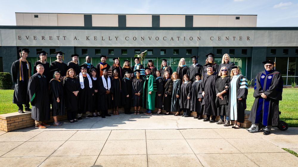 Graduates in academic regalia pose outside Everett A. Kelly Convocation Center during a university commencement ceremony.