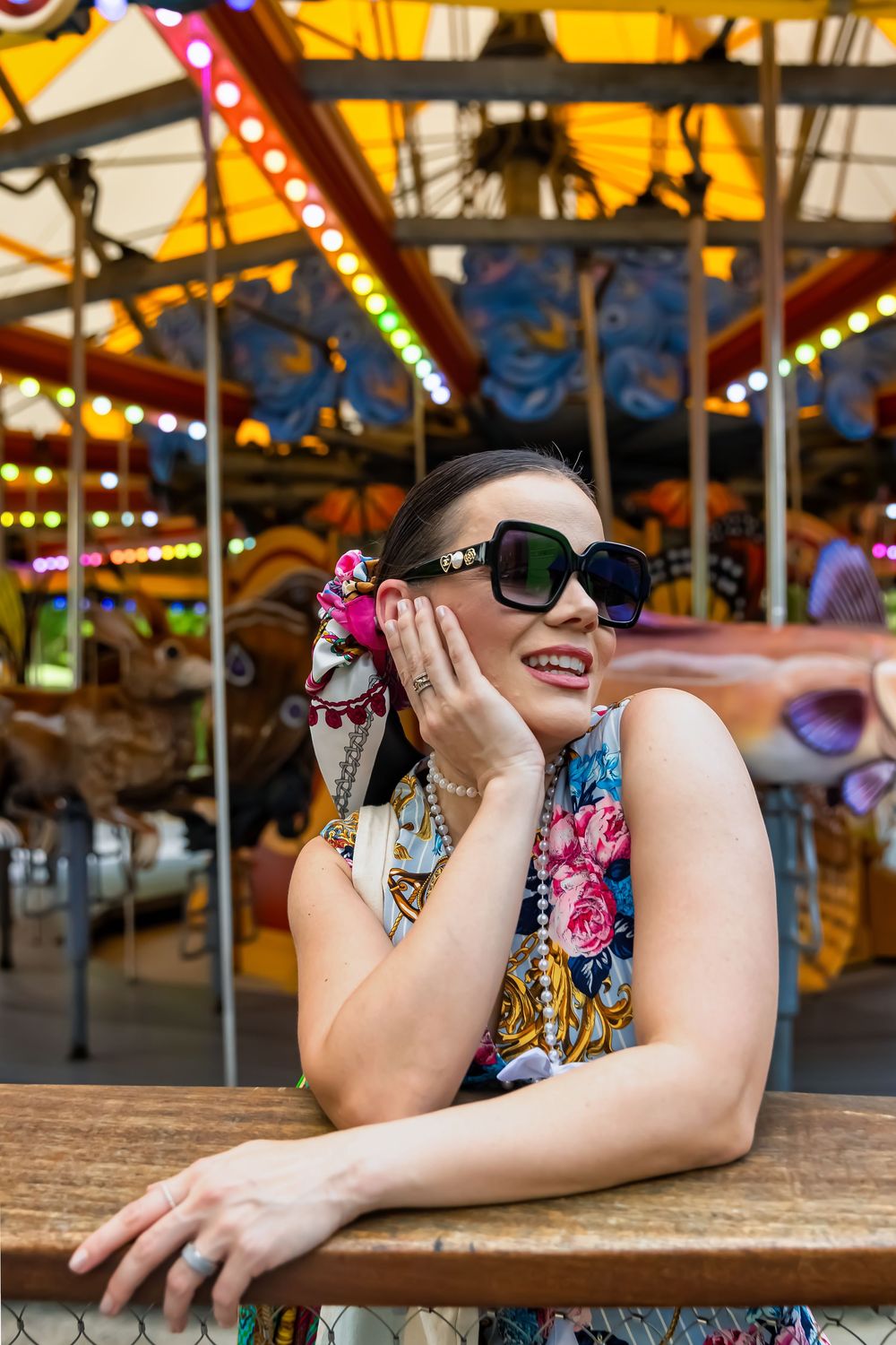 Woman in floral outfit and sunglasses poses by a colorful carousel at an amusement park.