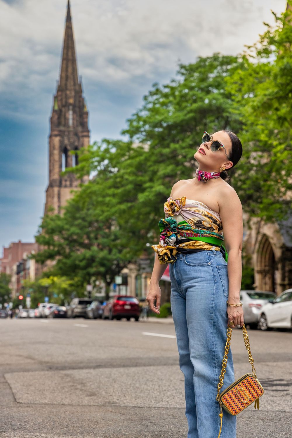 Stylish woman in colorful strapless top, wide-leg jeans, and sunglasses poses on urban street with church steeple backdrop.