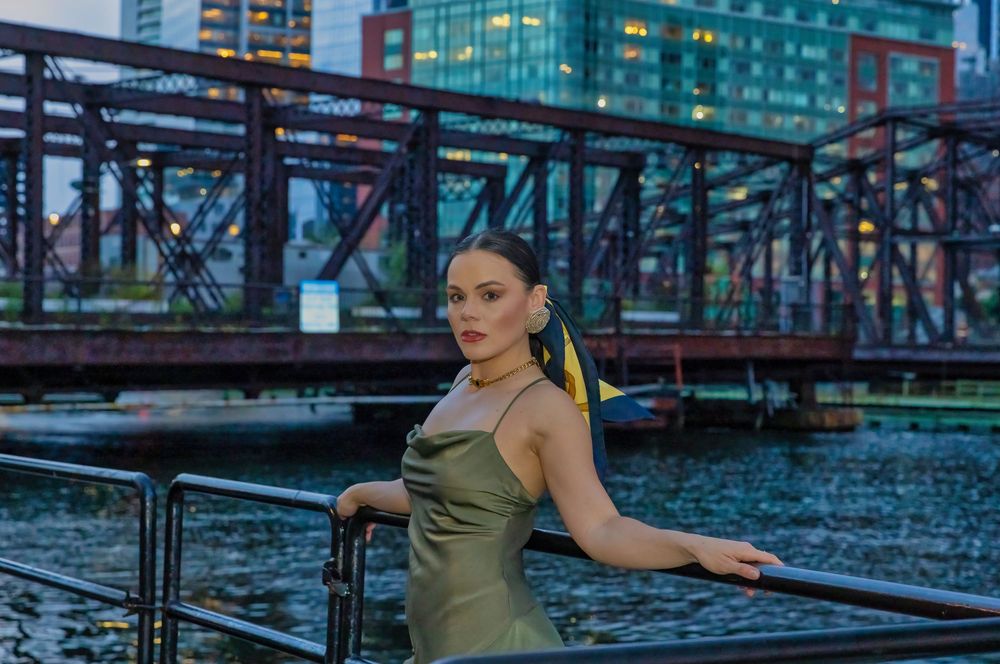 Young woman in olive dress poses by waterfront railing with blue steel bridge and city skyline at dusk.