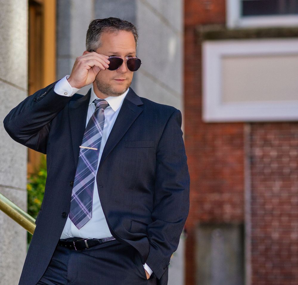 Man in navy suit adjusting sunglasses outdoors near brick building, professional business attire with plaid tie.
