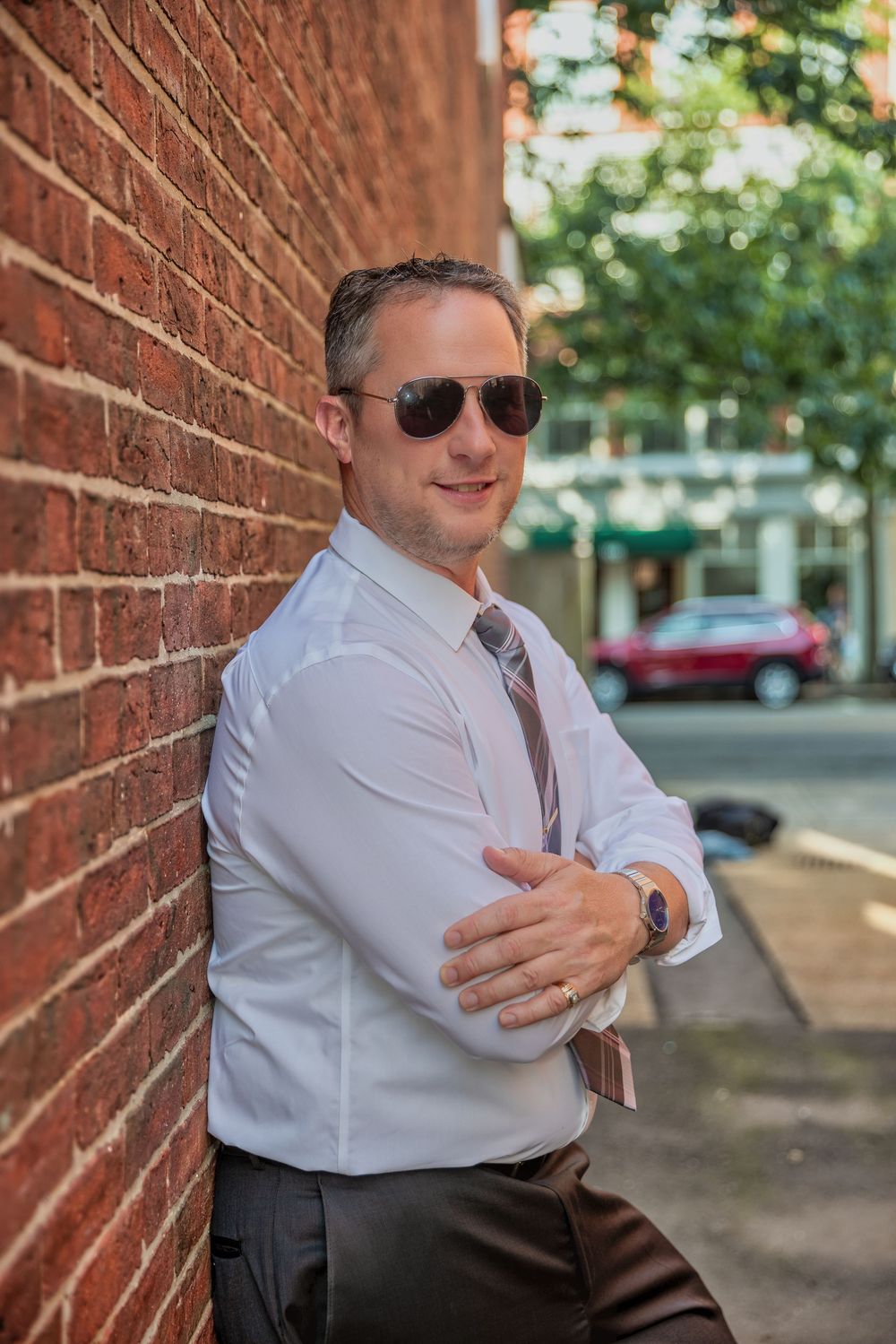 Man in white shirt, tie, and sunglasses leaning against red brick wall on urban street.