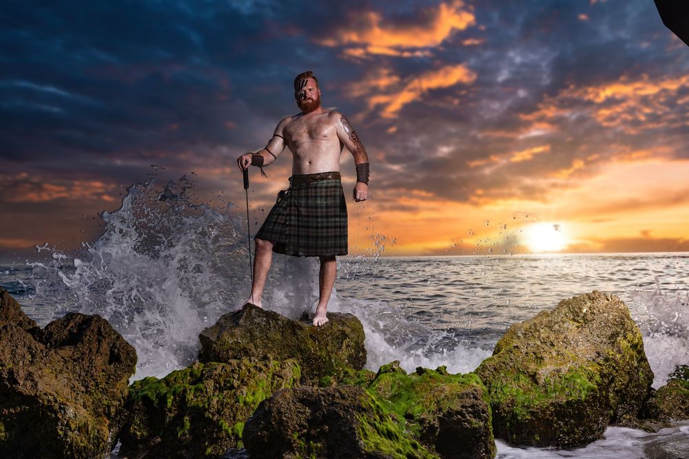 Shirtless man in tartan kilt stands on mossy rocks with ocean waves and dramatic sunset sky behind him.