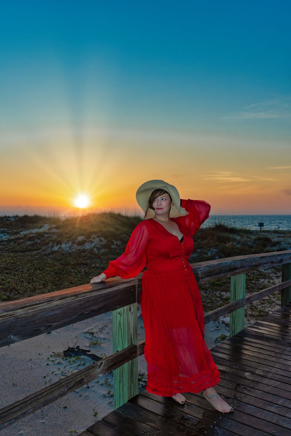 Woman in red dress and hat poses on beach boardwalk at sunset with golden rays over sand dunes.