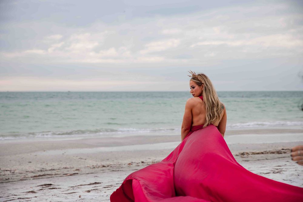 Woman in flowing pink gown poses on sandy beach with ocean waves and cloudy sky backdrop.