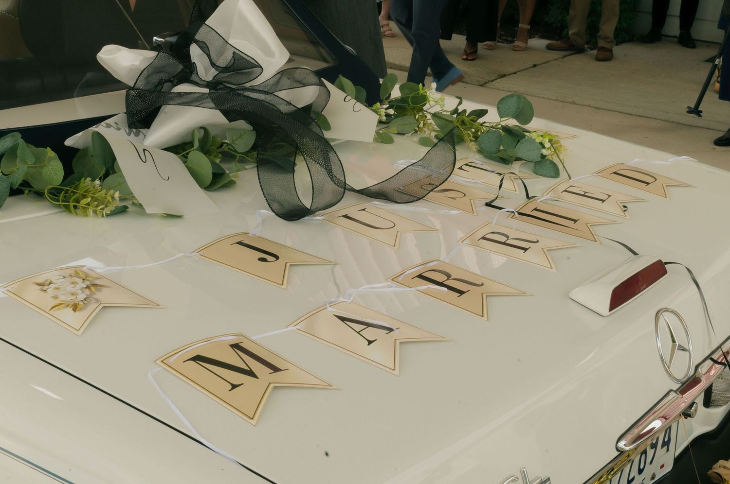 White Mercedes wedding car decorated with floral banner spelling MARRY, black organza bow, and greenery eucalyptus accents.
