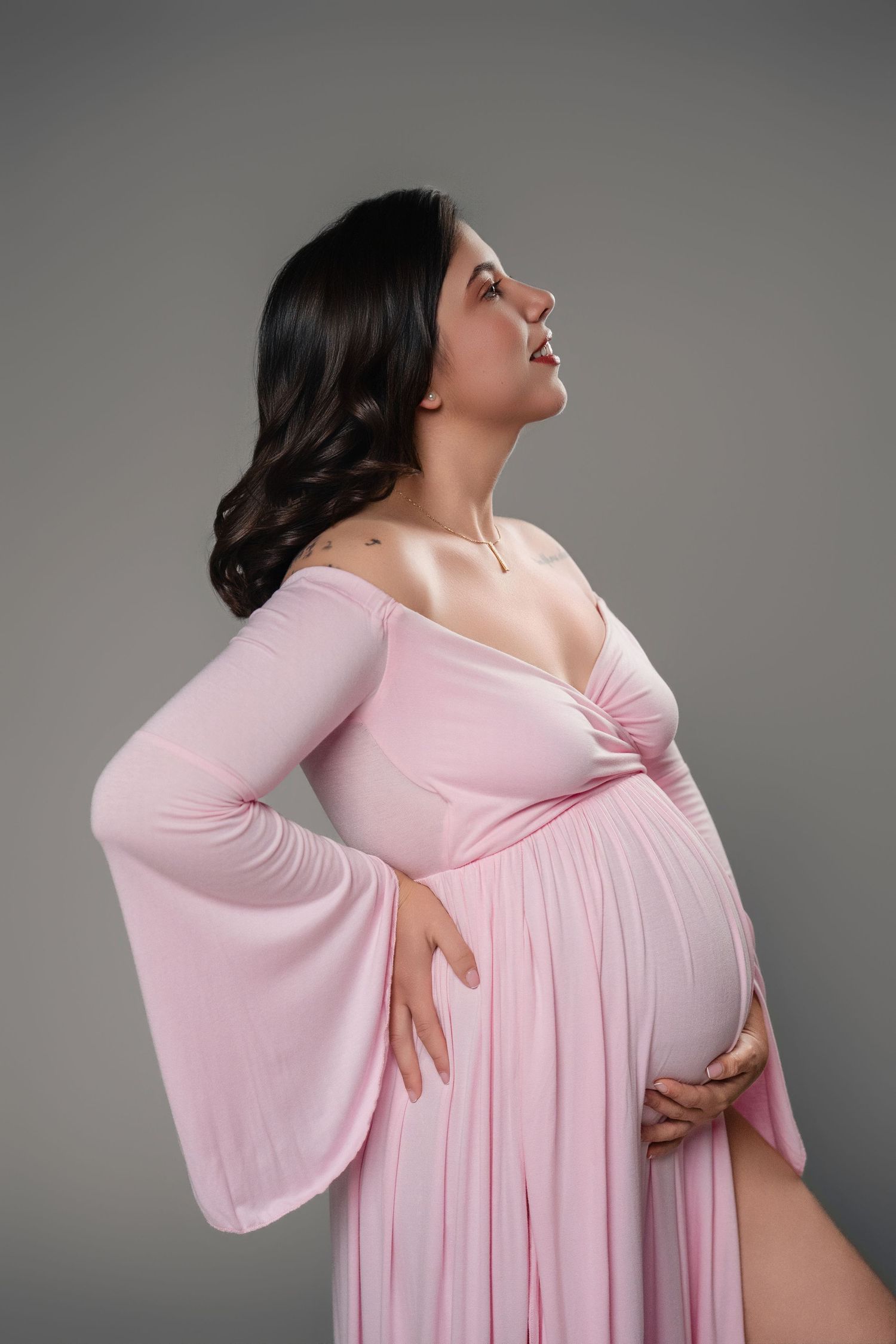 Pregnant woman in pink off-shoulder gown cradling baby bump, studio maternity portrait on gray background.