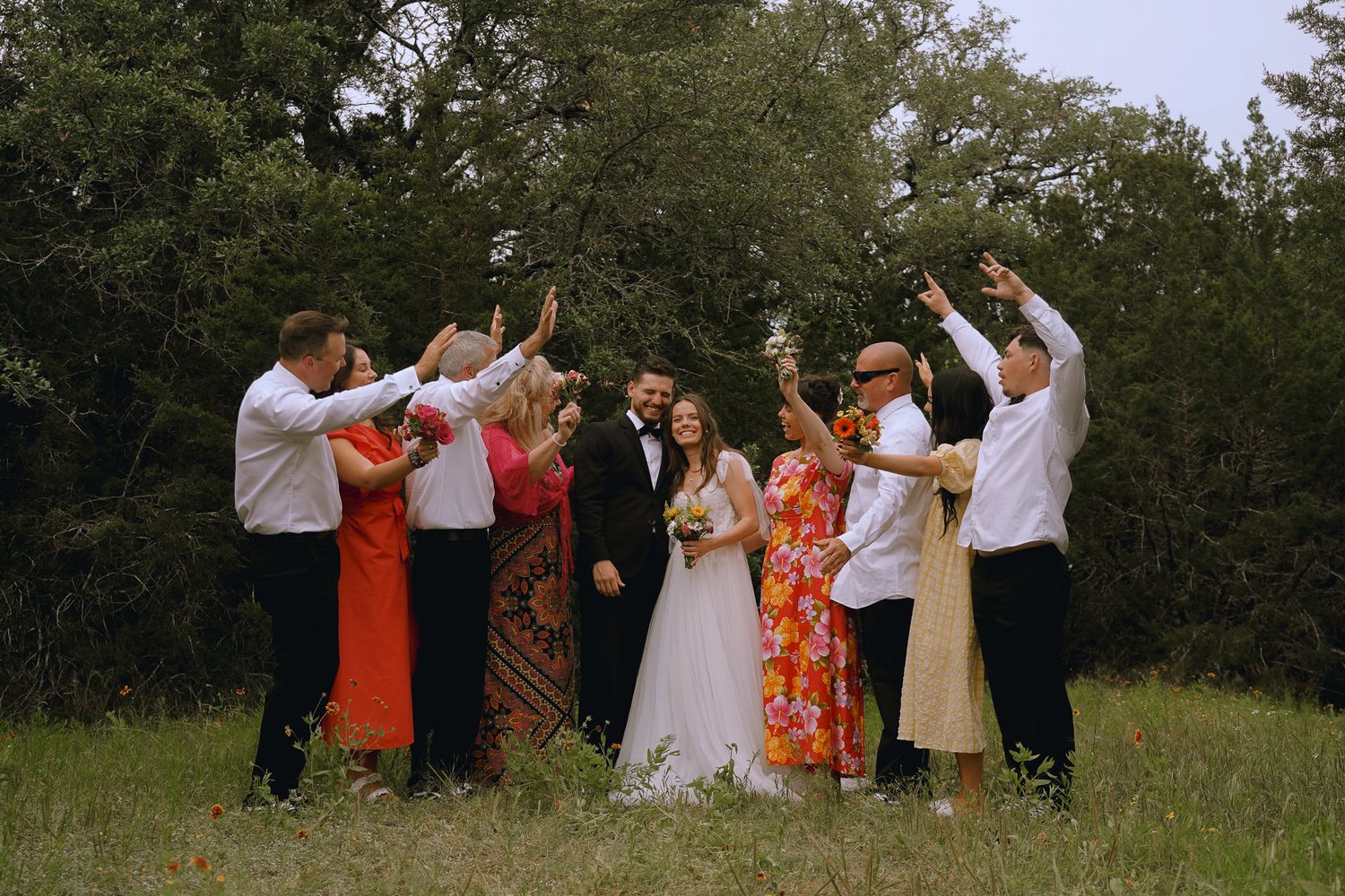 Wedding party celebrating outdoors with raised hands, bride in white gown, guests in colorful attire, lush green trees background.