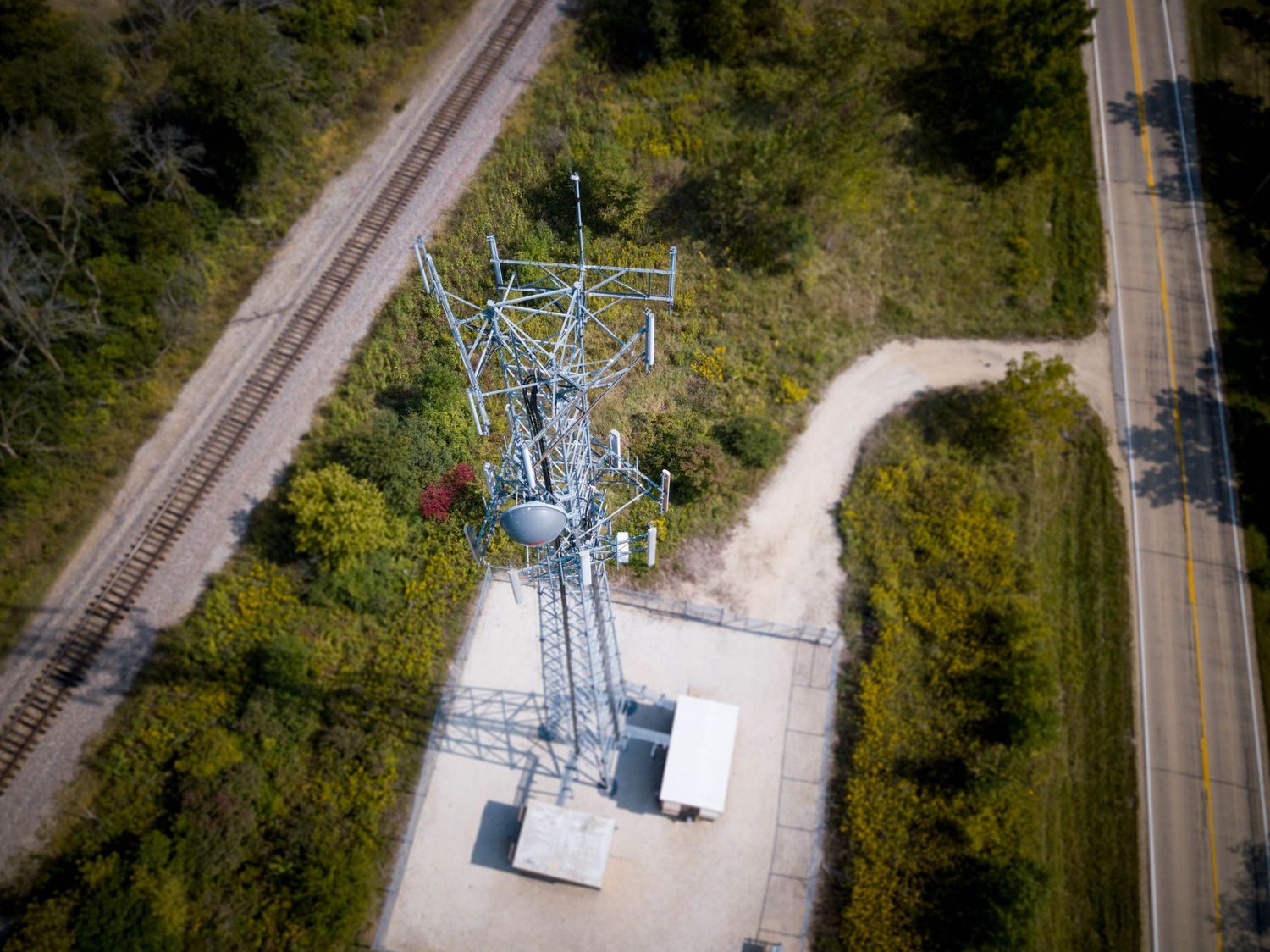 Aerial view of an electrical substation tower near railroad tracks and a rural road surrounded by trees.