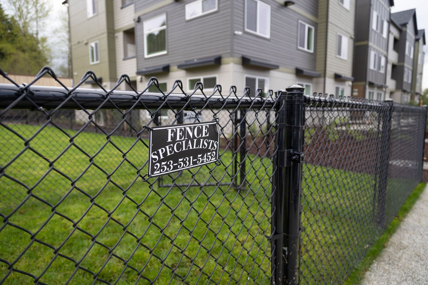 Black chain-link fence with Fence Specialists company sign installed near modern apartment complex.