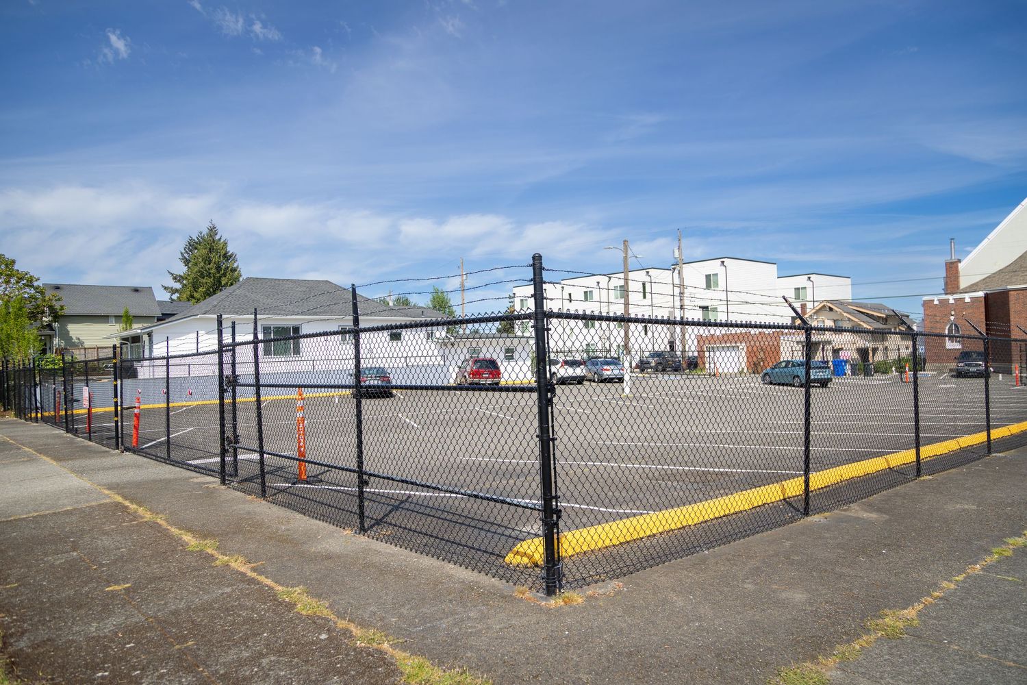 Black chain-link fenced parking lot with yellow markings near commercial buildings under a clear blue sky.