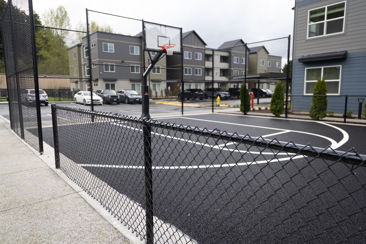 Outdoor basketball court with chain-link fence surrounded by modern apartment buildings and parked cars.