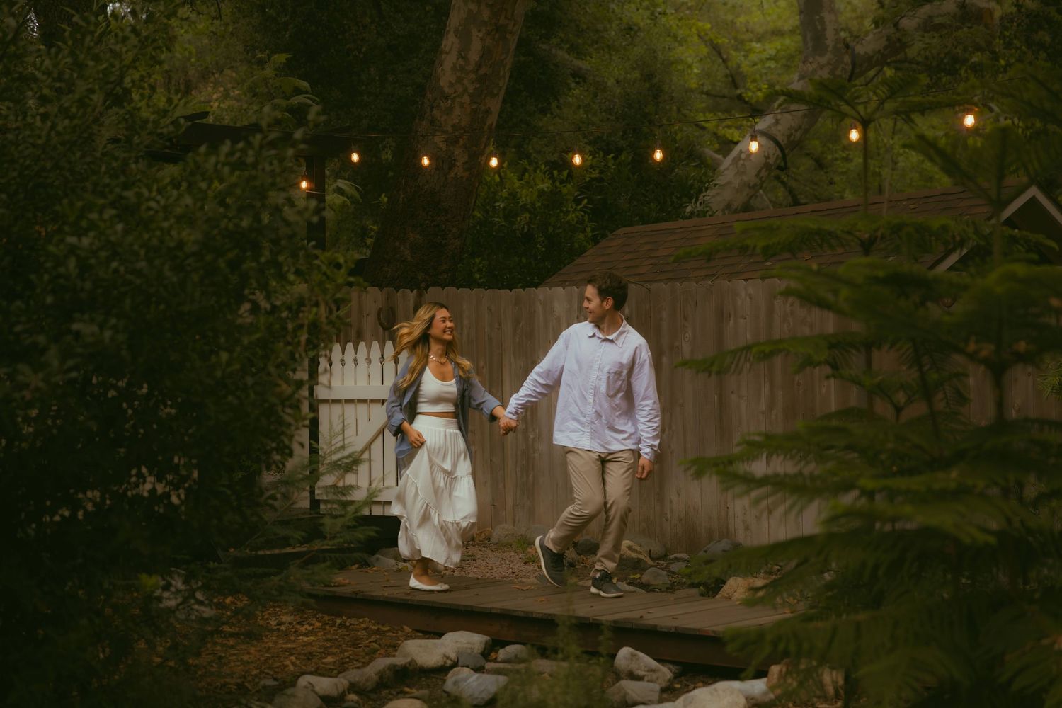 Couple holding hands walking on a wooden deck surrounded by lush greenery and warm string lights at dusk.
