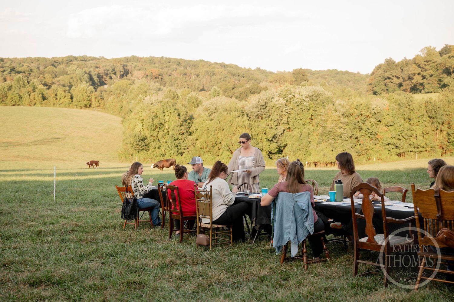 Outdoor dinner party with guests seated at black-draped tables on a green lawn, surrounded by lush trees at golden hour.