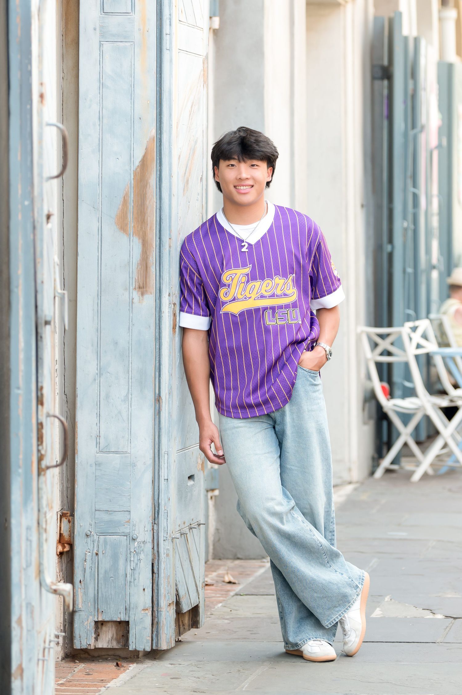 Young man in purple Tigers jersey and wide-leg jeans leaning against weathered door on city sidewalk.
