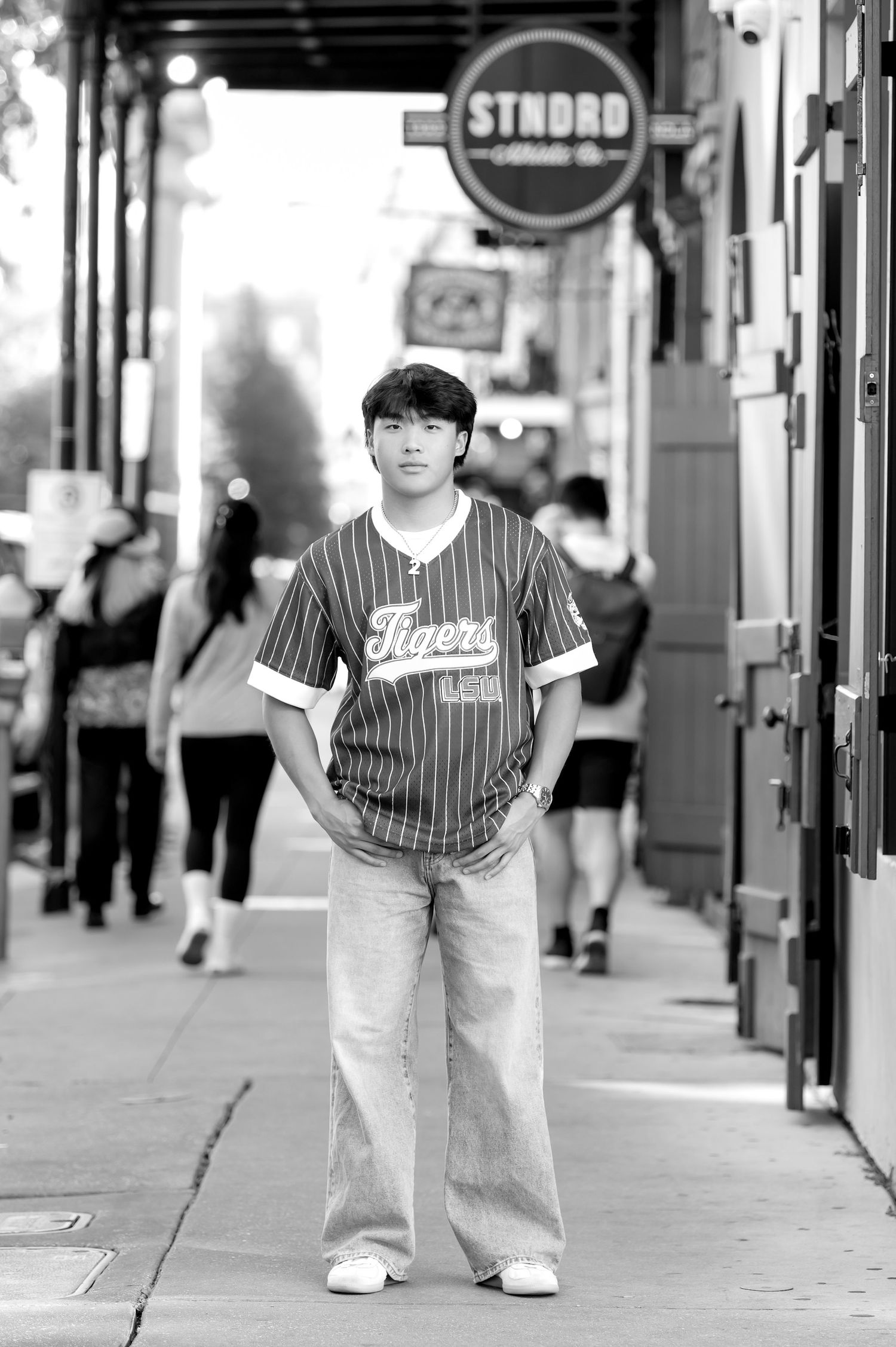 Young man in Tigers baseball jersey and wide-leg jeans stands on urban sidewalk in black and white photo.