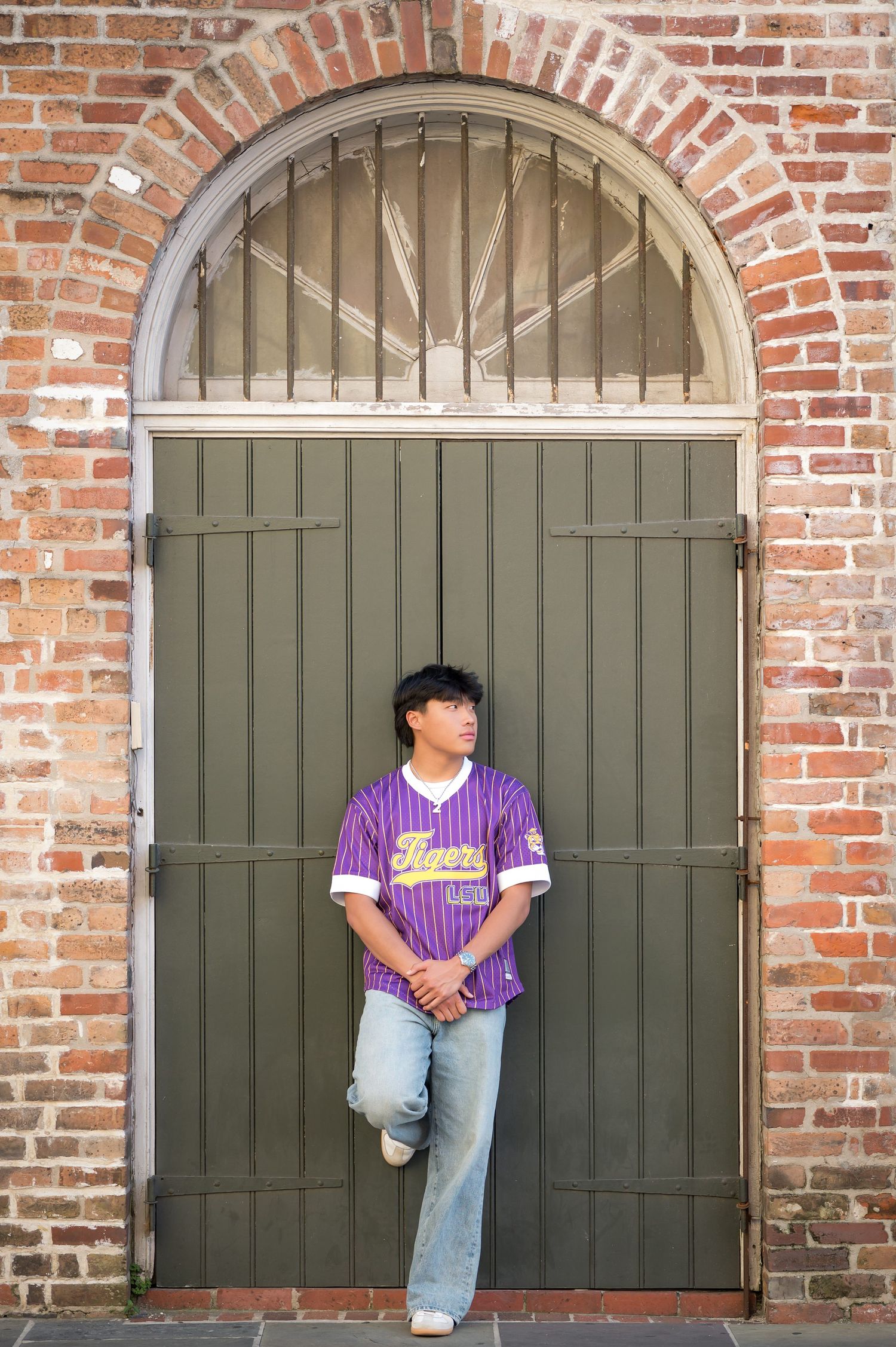 Young man in purple pinstripe baseball jersey leans against ornate green door in brick archway.