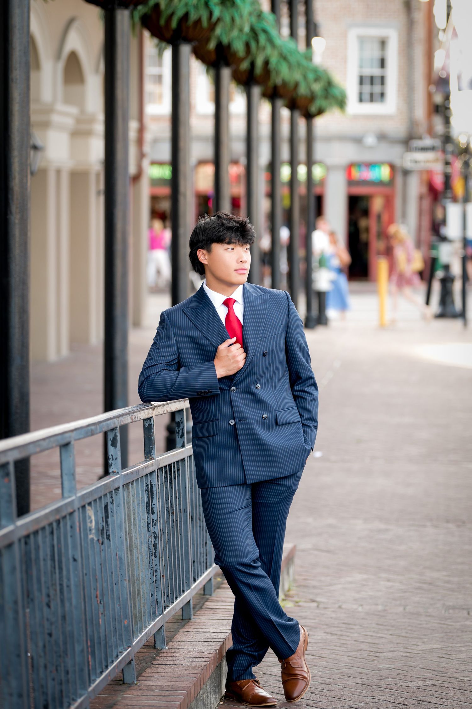 Young man in navy blue suit and red tie leaning against railing on urban sidewalk.