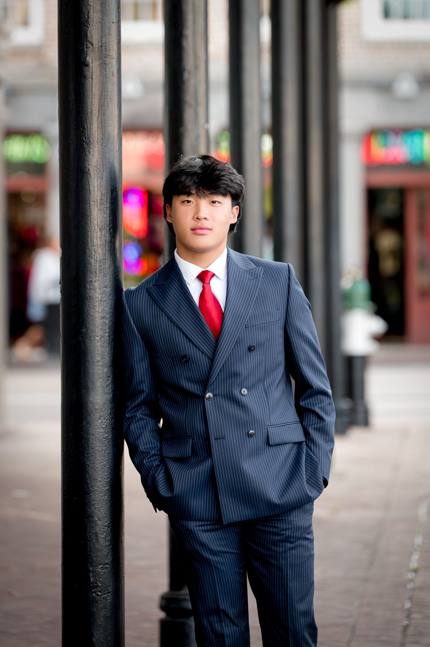 Young Asian man in navy pinstripe double-breasted suit and red tie leaning against black pole on city street.