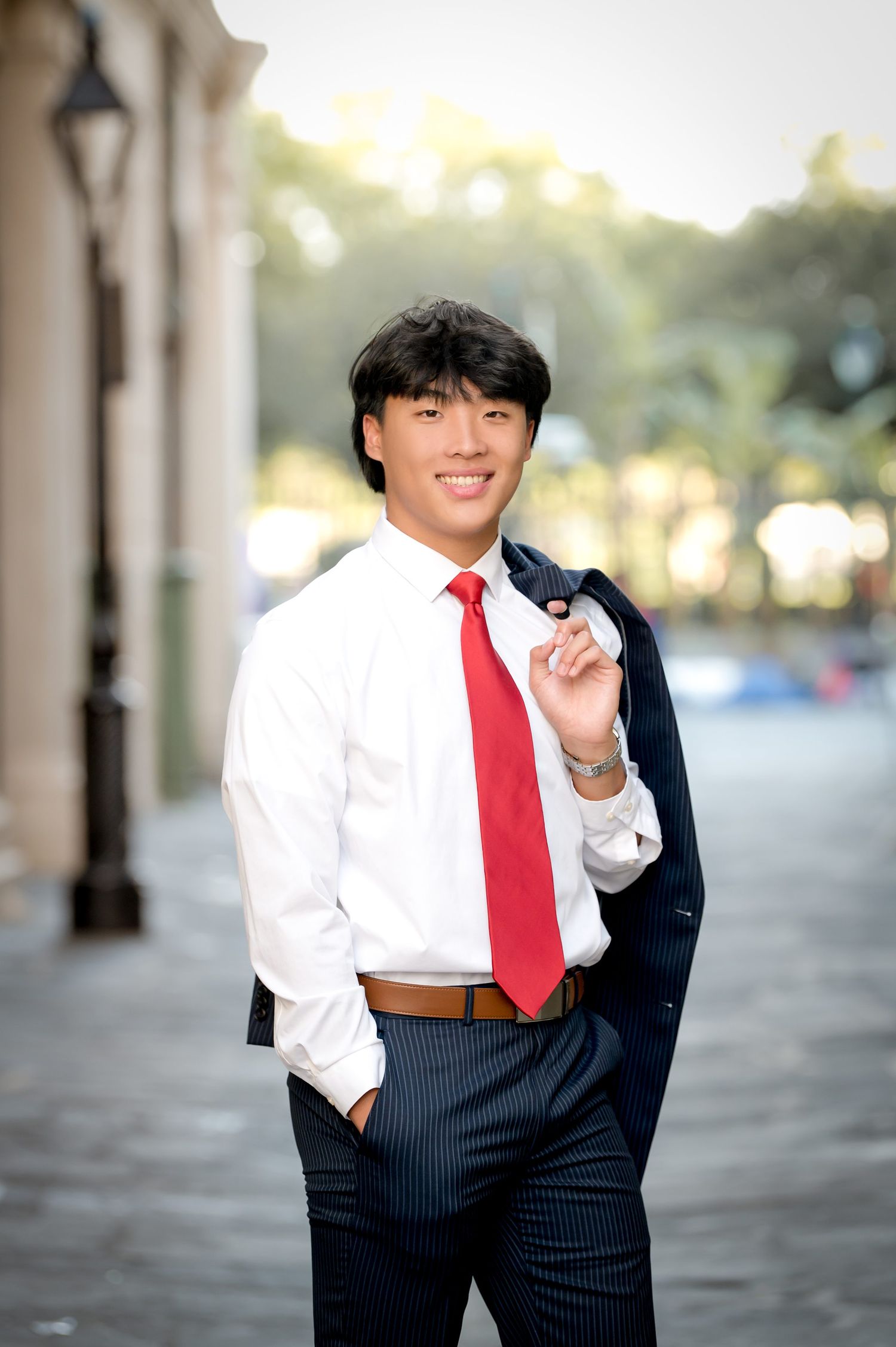 Smiling young man in white shirt and red tie holds suit jacket over shoulder on cobblestone street.