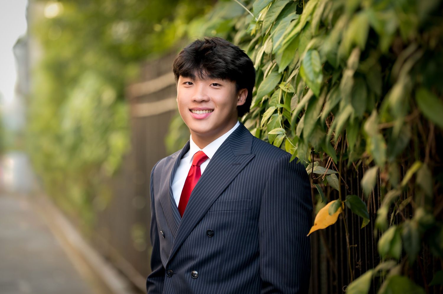 Young Asian man in pinstripe suit and red tie smiling outdoors against lush green ivy-covered wall.