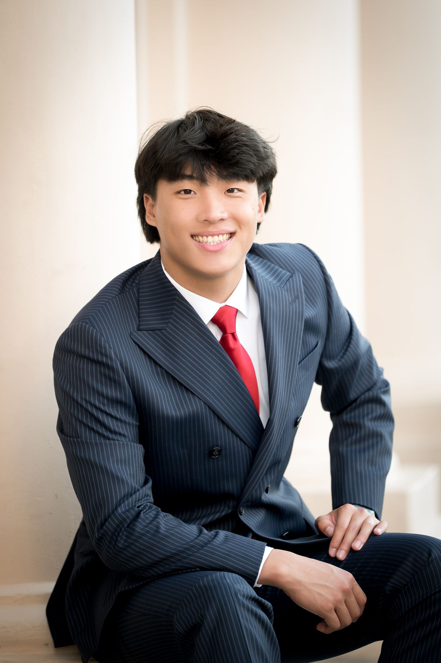 Young man in pinstripe suit and red tie smiling confidently while seated in a professional portrait setting.