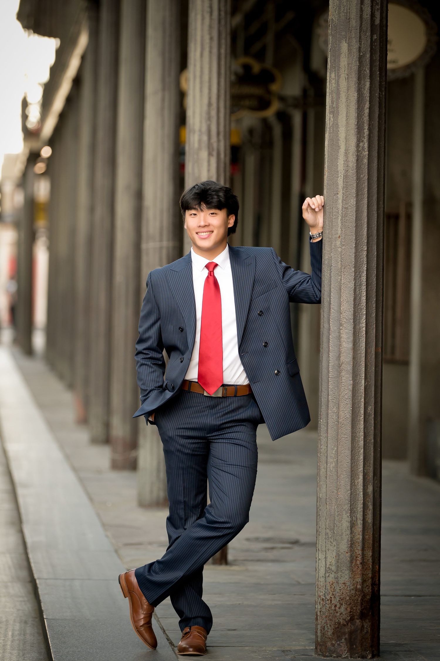 Young Asian man in navy suit and red tie smiling while leaning against a stone column on a city sidewalk.