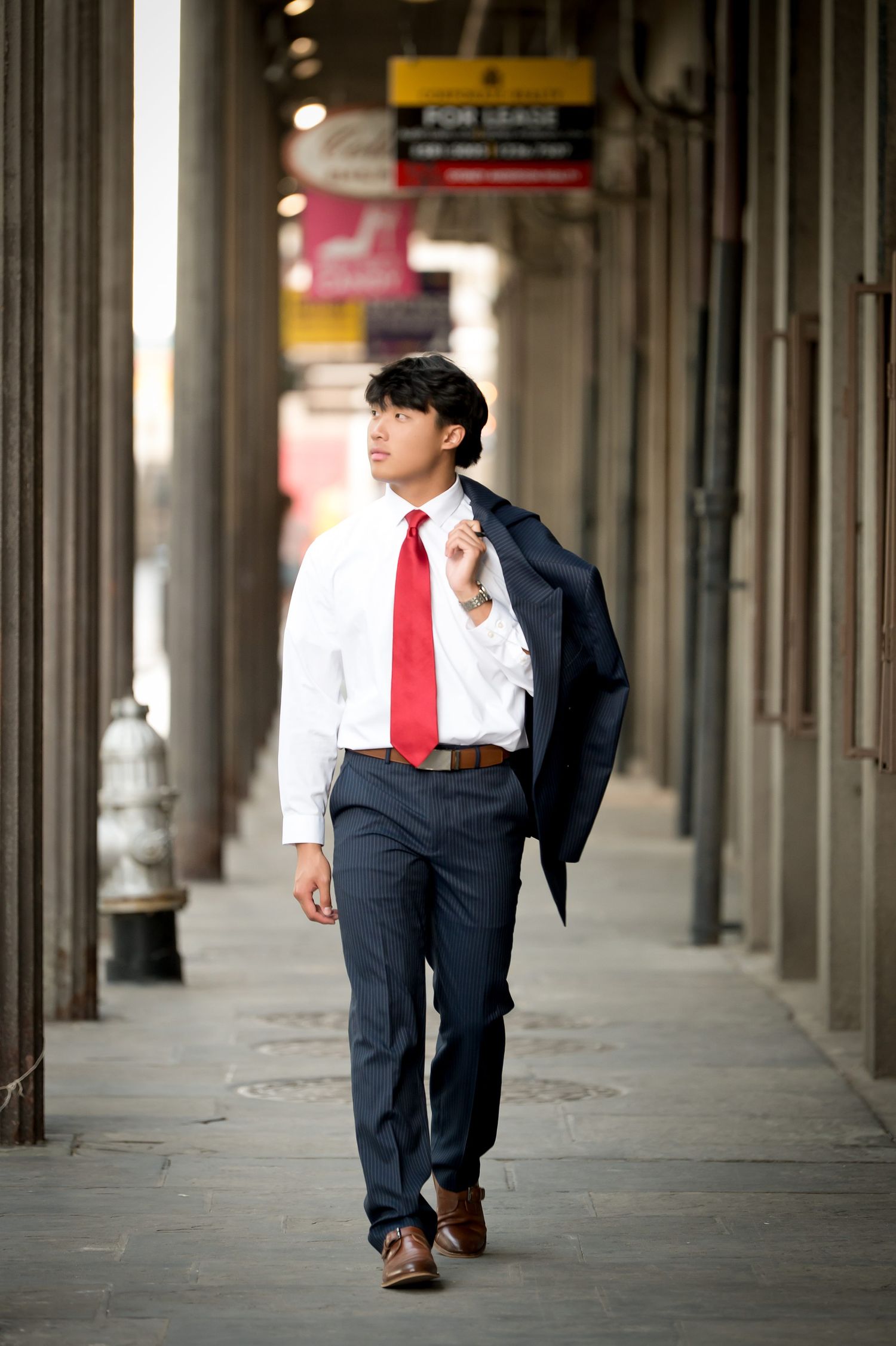 Young Asian man walking under urban colonnade in white shirt and red tie carrying navy suit jacket over shoulder.