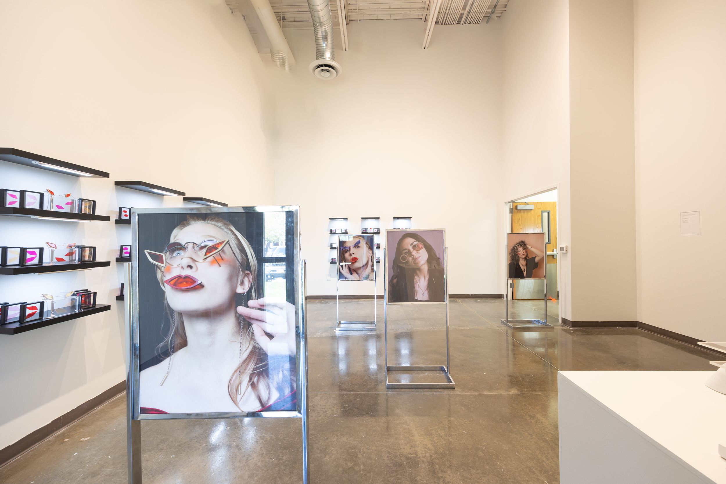 Gallery space displaying large portrait photographs on freestanding panels with shelving.
