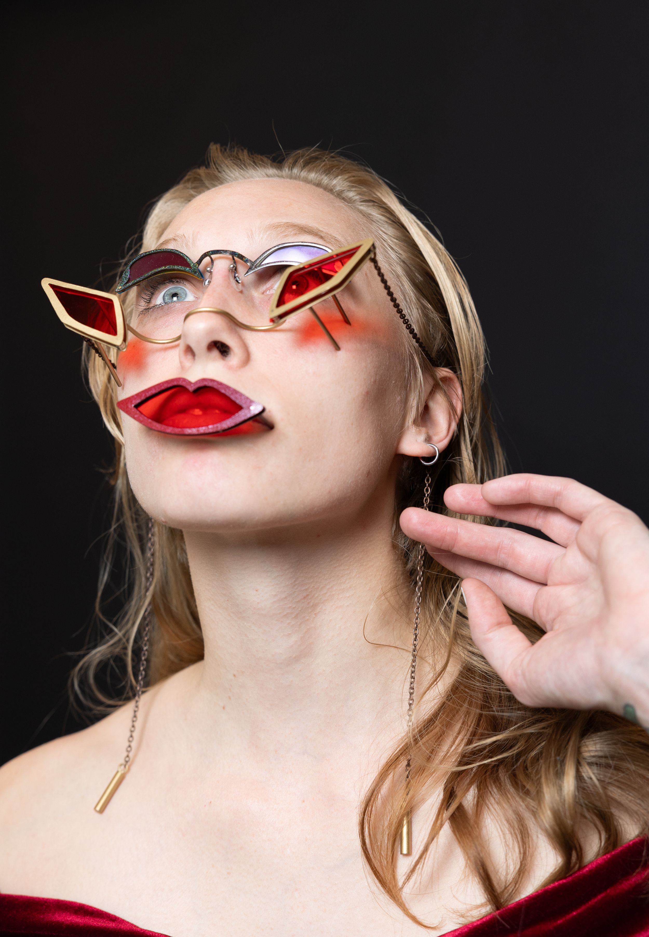 Woman wearing avant-garde decorative glasses with red lip design against a dark studio background.