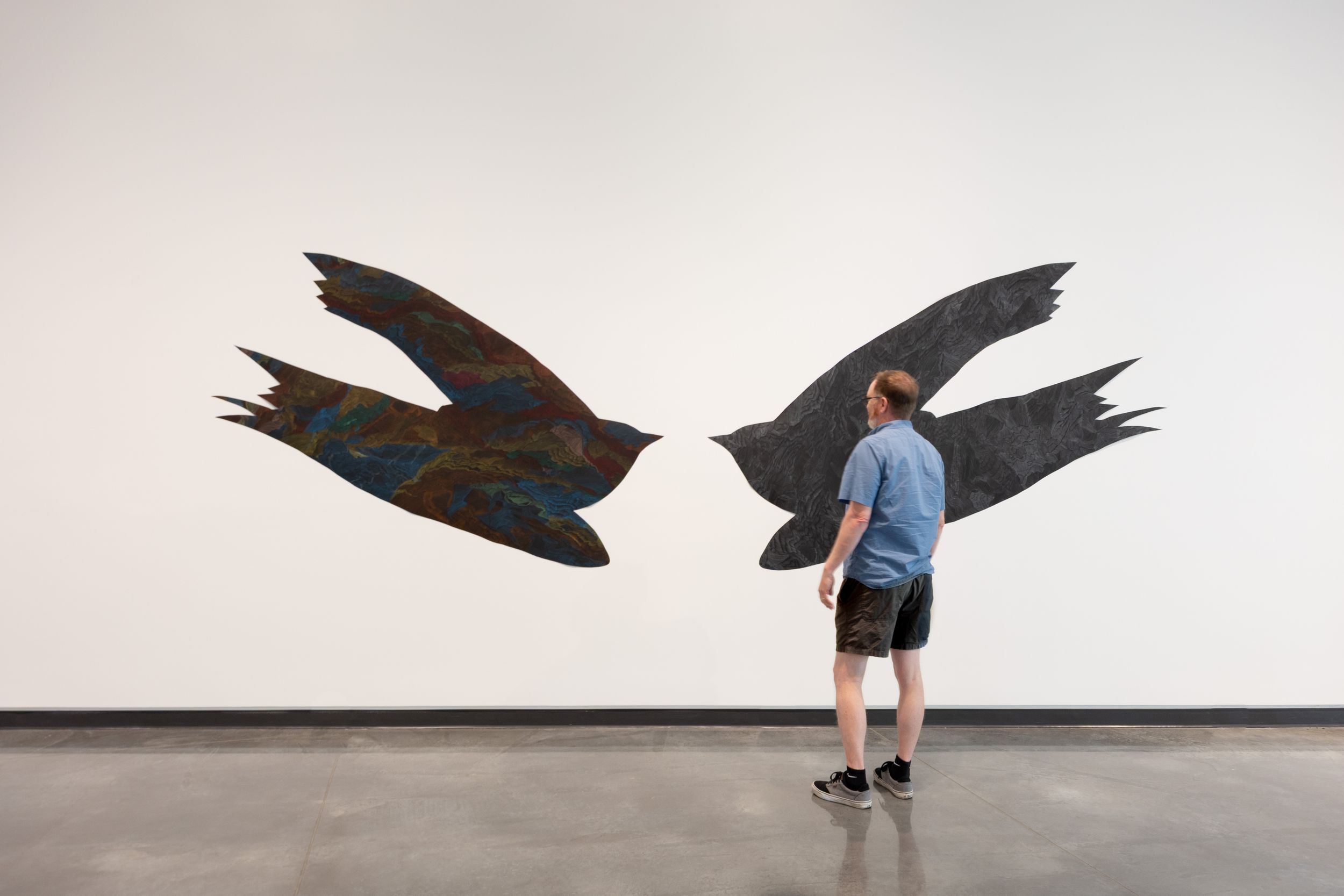 A man stands before a large wall mural of two dark birds facing each other in a gallery.