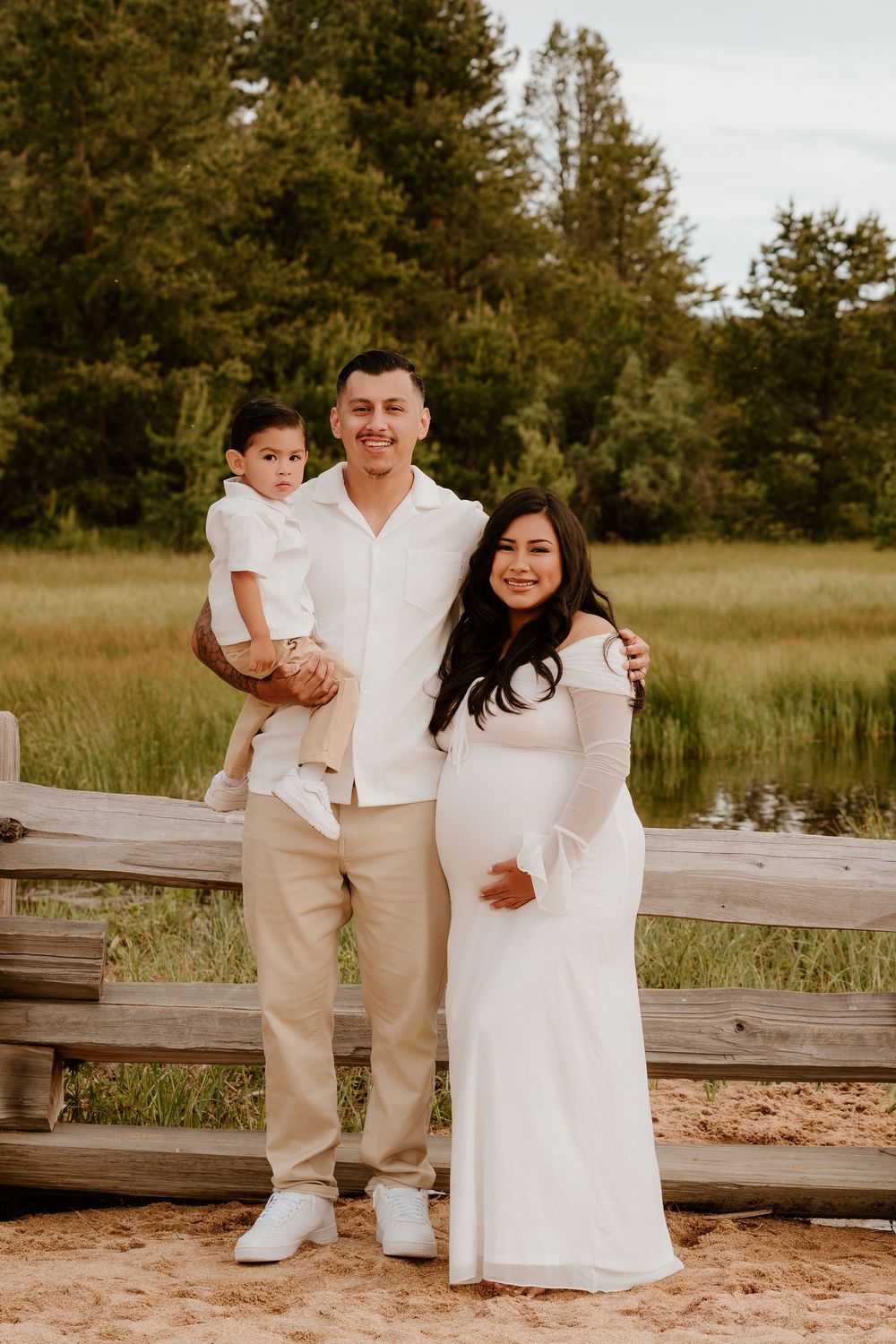 Family maternity photo with couple and toddler in white outfits near scenic mountain meadow.
