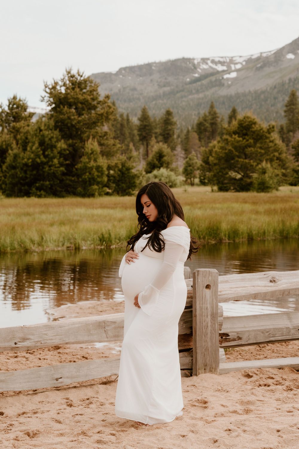 Pregnant woman in white off-shoulder gown cradling baby bump near mountain lake with pine trees.