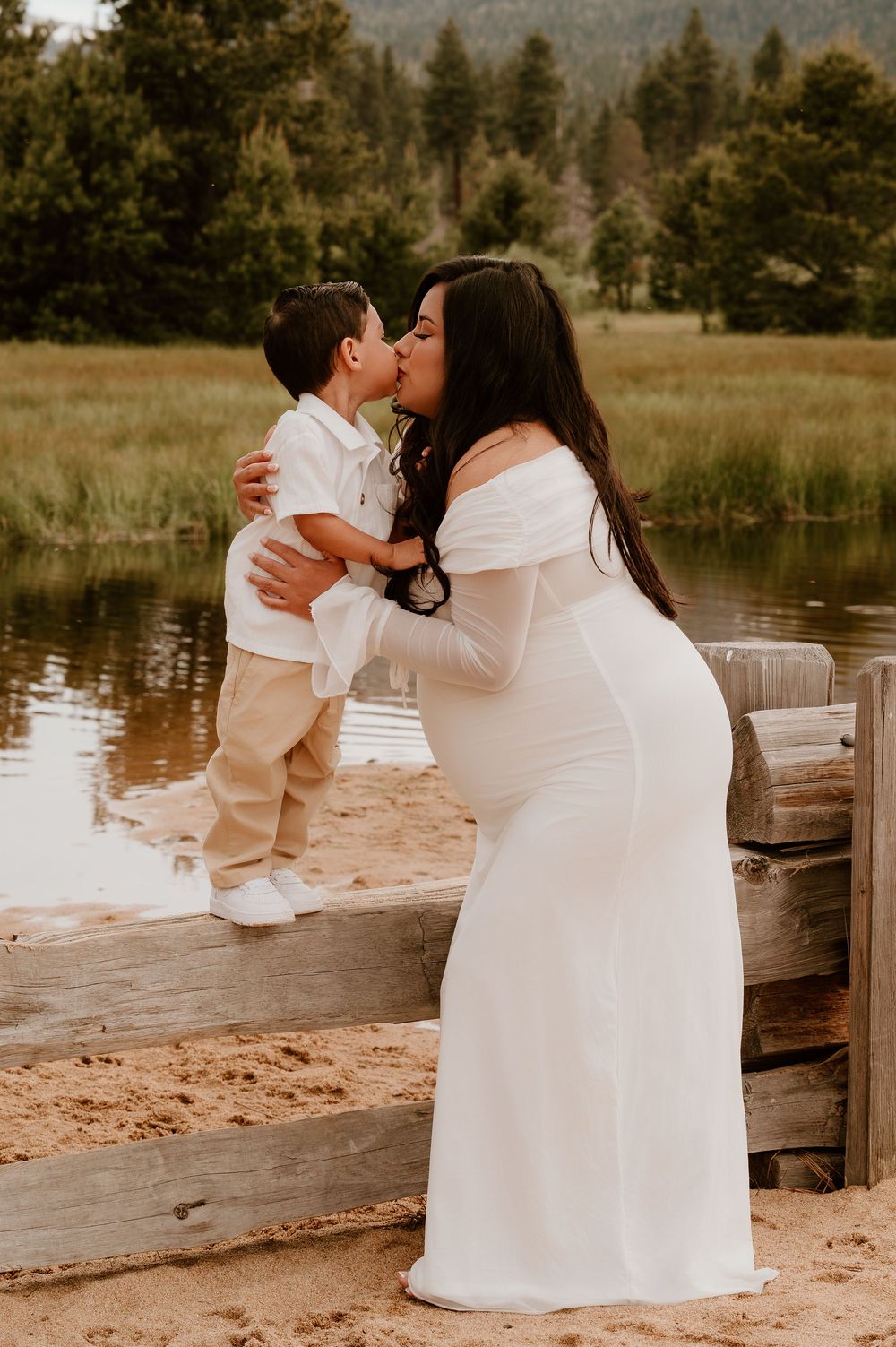 Pregnant woman in white gown kisses young boy outdoors near a pond with green meadows and pine trees.
