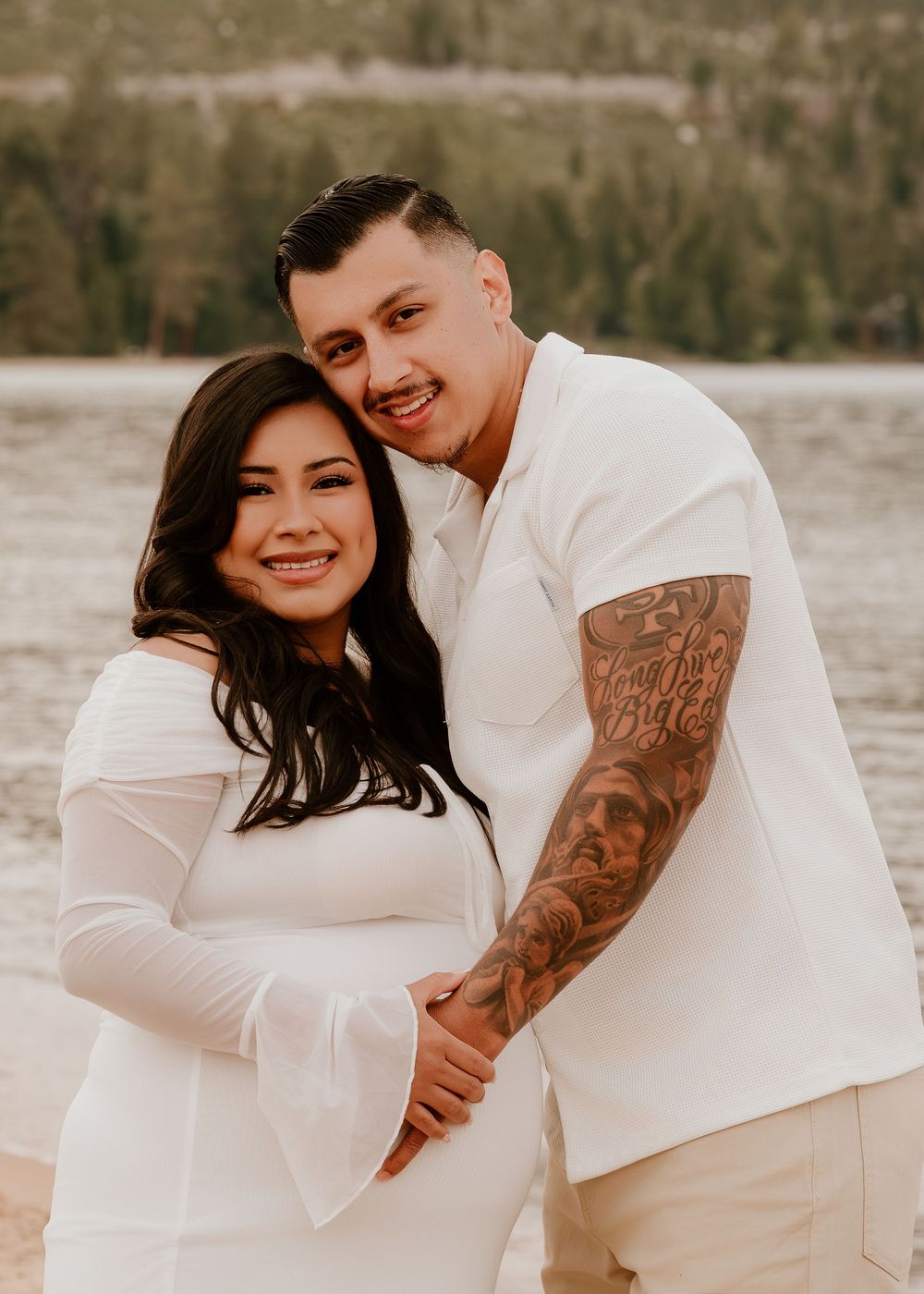 Smiling couple in matching white outfits pose together near a lake, woman showing baby bump in a maternity photo shoot.
