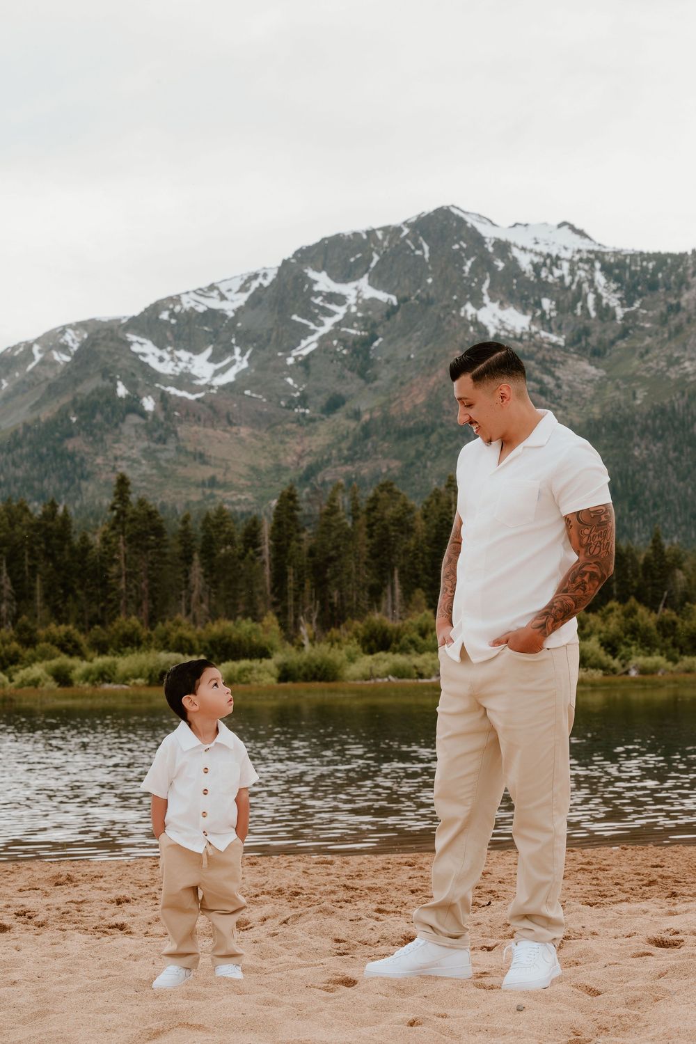 Father and young son in matching white outfits stand on sandy beach with mountain lake and snow-capped peaks backdrop.