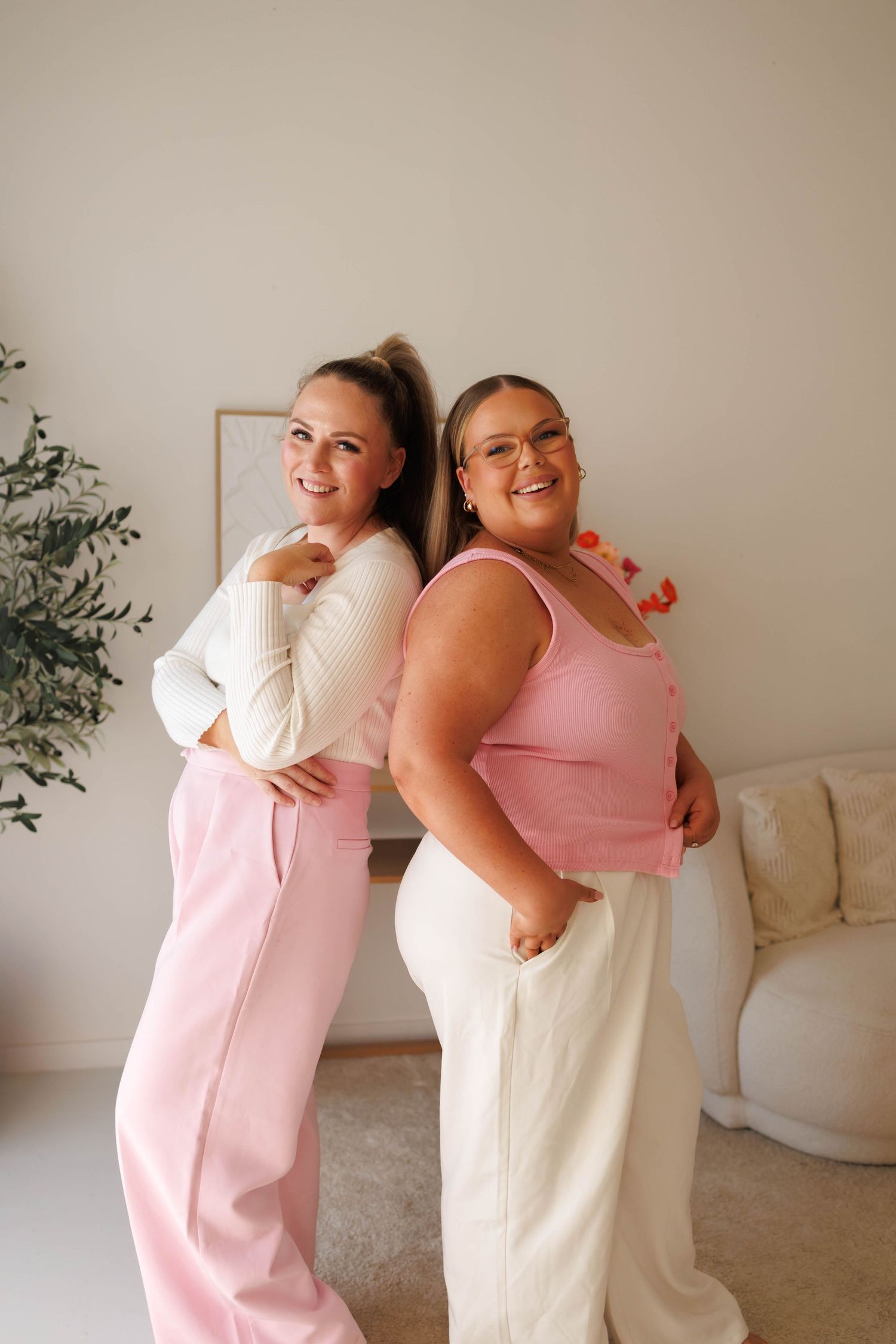 Two smiling women posing back-to-back in pink and white outfits in a bright, minimalist living room.