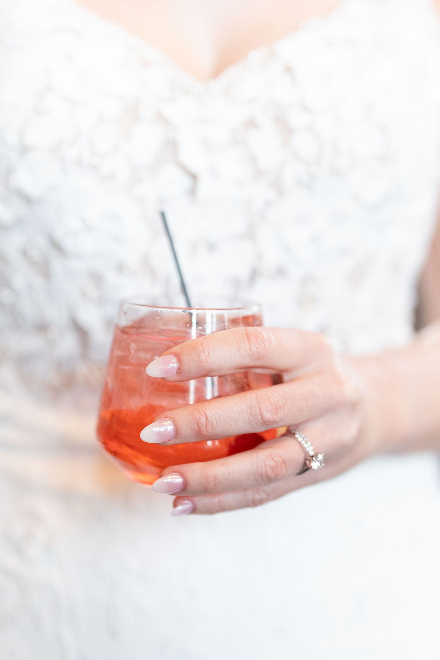 Bride in white lace dress holding a stemless glass with a pink cocktail and straw, wearing a diamond ring.