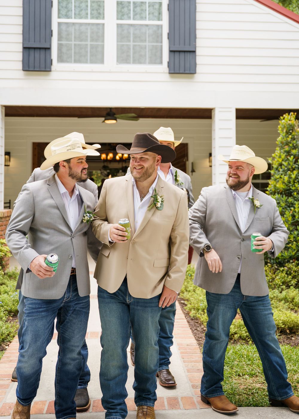 Four groomsmen in cowboy hats, gray suits, and jeans holding drinks outside a white farmhouse wedding venue.