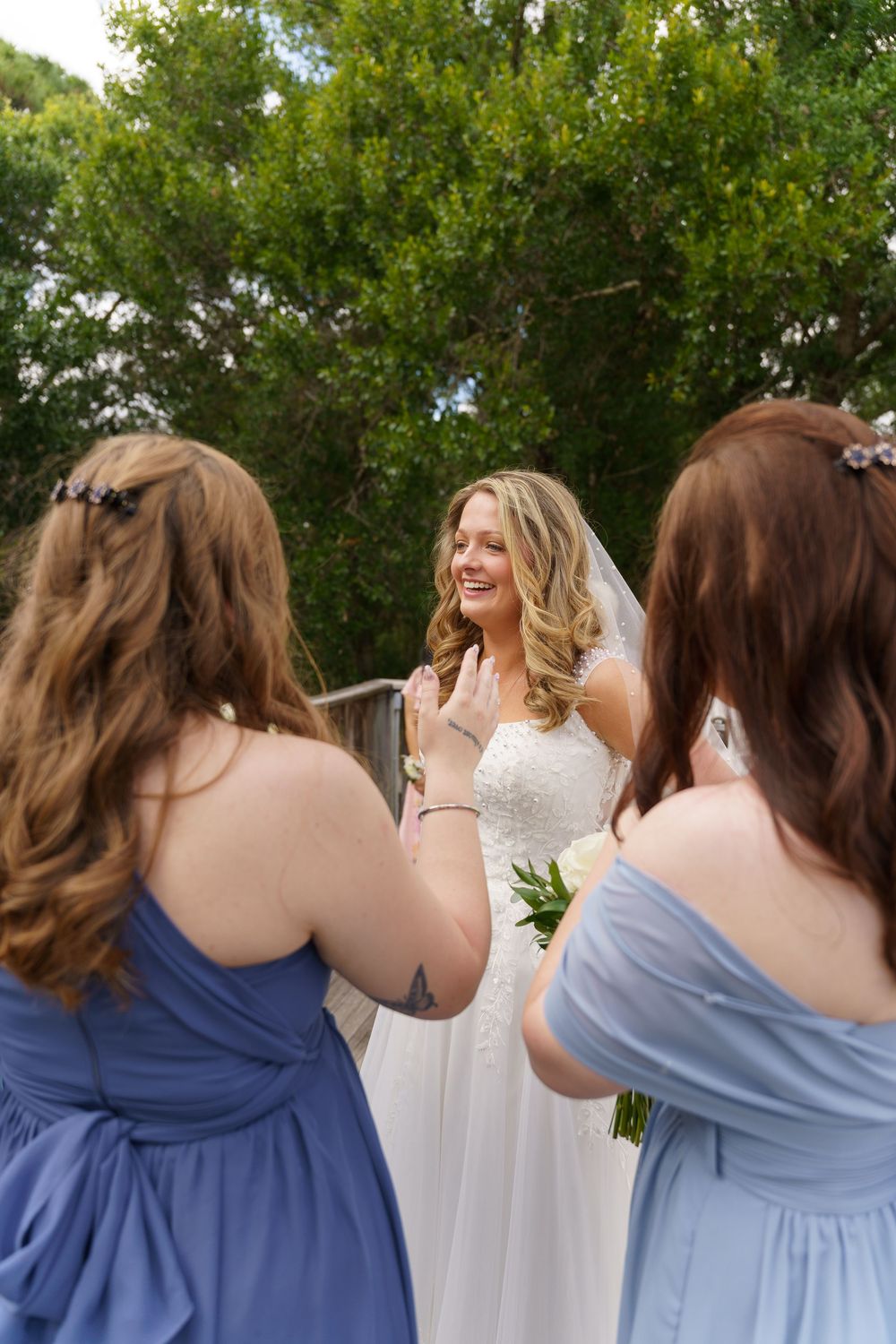 Bride in white gown and veil smiles emotionally at two bridesmaids in blue dresses outdoors near lush green trees.
