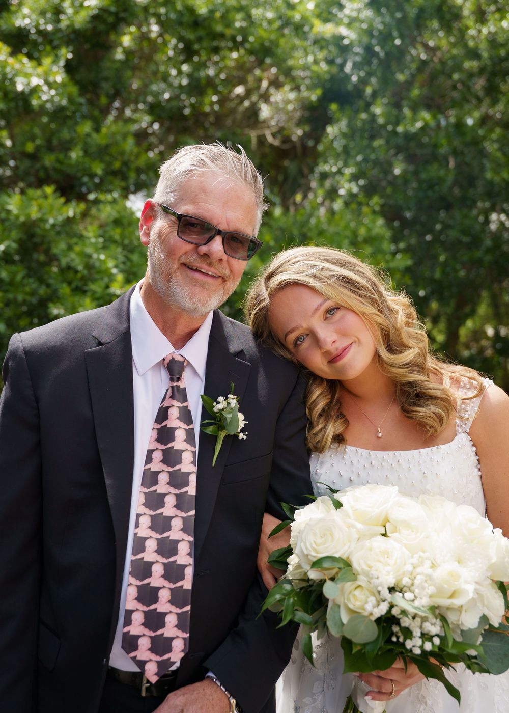 Father and bride pose together outdoors, she holds white roses bouquet, wearing wedding dress, he wears dark suit with patterned tie.
