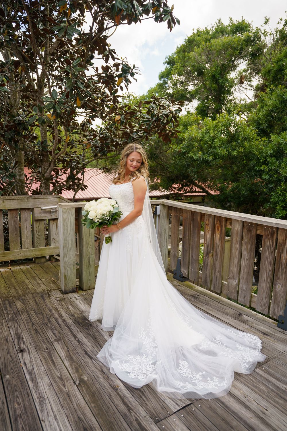 Bride in white lace wedding gown holding bouquet on wooden deck surrounded by lush greenery.