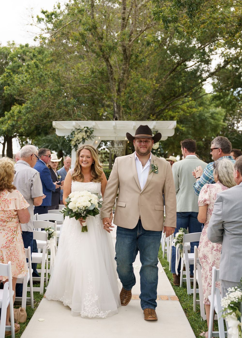 Bride and groom walk down outdoor wedding aisle, she holds white bouquet, he wears tan blazer and cowboy hat.