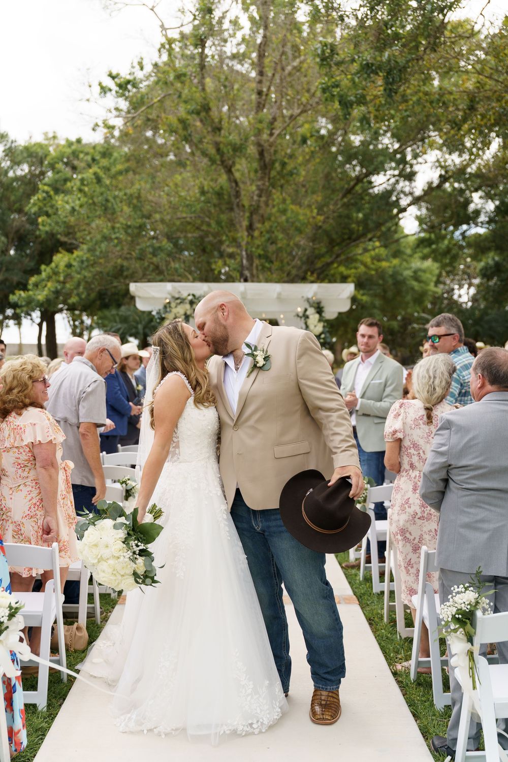 Bride and groom share a kiss at the end of the aisle after outdoor wedding ceremony, groom in tan blazer and jeans.
